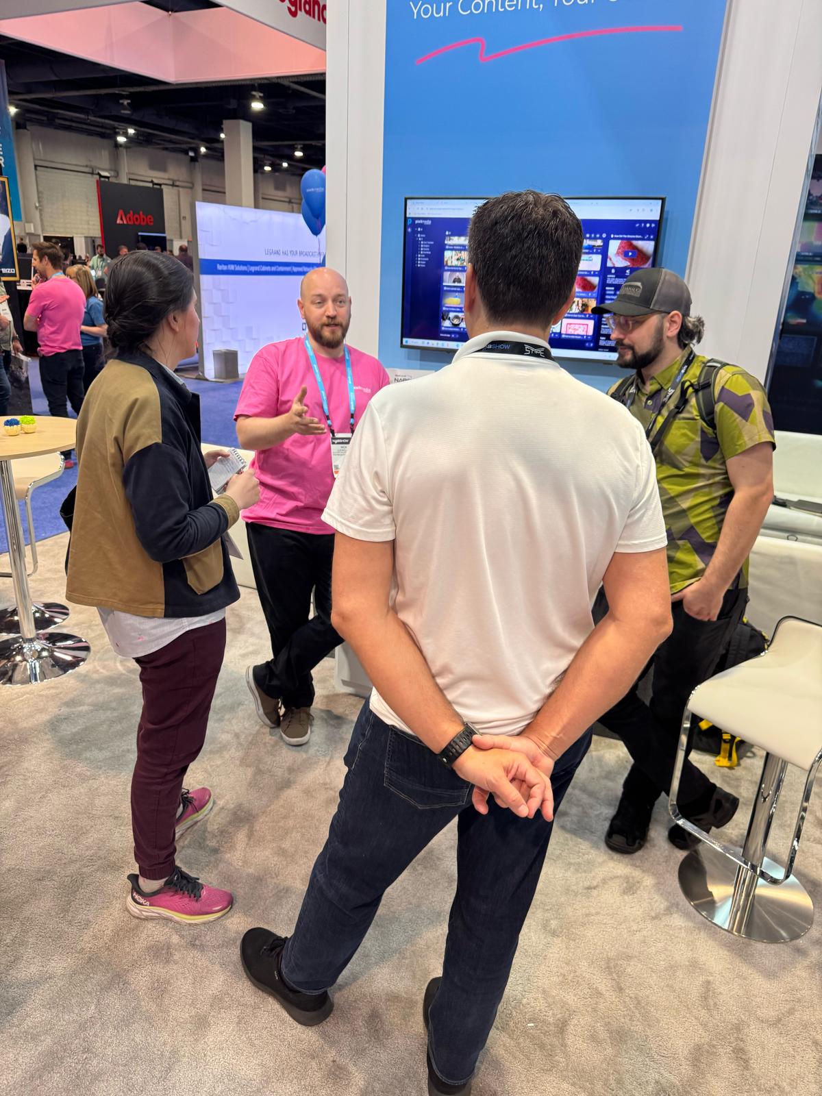 Engaging three visitors in a brightly lit exhibition hall. Conversing at a Pixitmedia booth. The booth highlights media management with a demo screen display.
