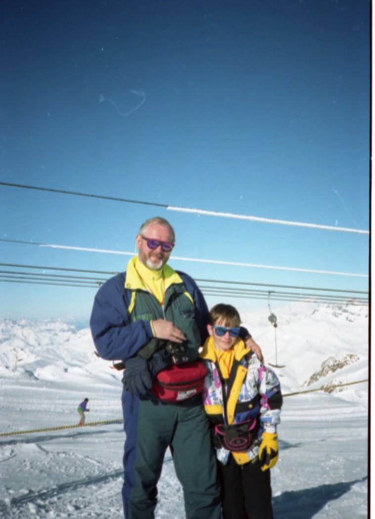 A father and young son standing in snow against a mountain backdrop at Les Deux Alpes ski resort. Both are wearing colorful ski gear and sunglasses, with ski lift cables visible overhead against a bright blue sky.