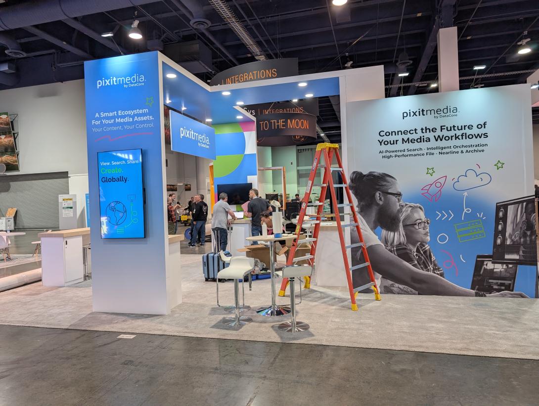 The calm before the storm. Pixitmedia booth setup at a pre-NAB show. Blue and white branded panels highlight media workflow solutions. An orange ladder indicates ongoing assembly.