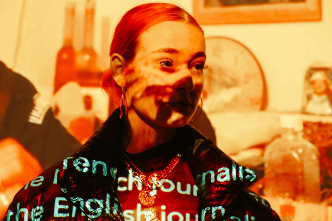 A young woman with reddish hair pulled back, wearing large gold hoop earrings and a gold chain necklace, looks off to the right. Text is projected onto her face and clothing, with words like "French," "English," and "our" partially visible. The background is blurred but suggests a domestic setting with bottles and framed pictures.