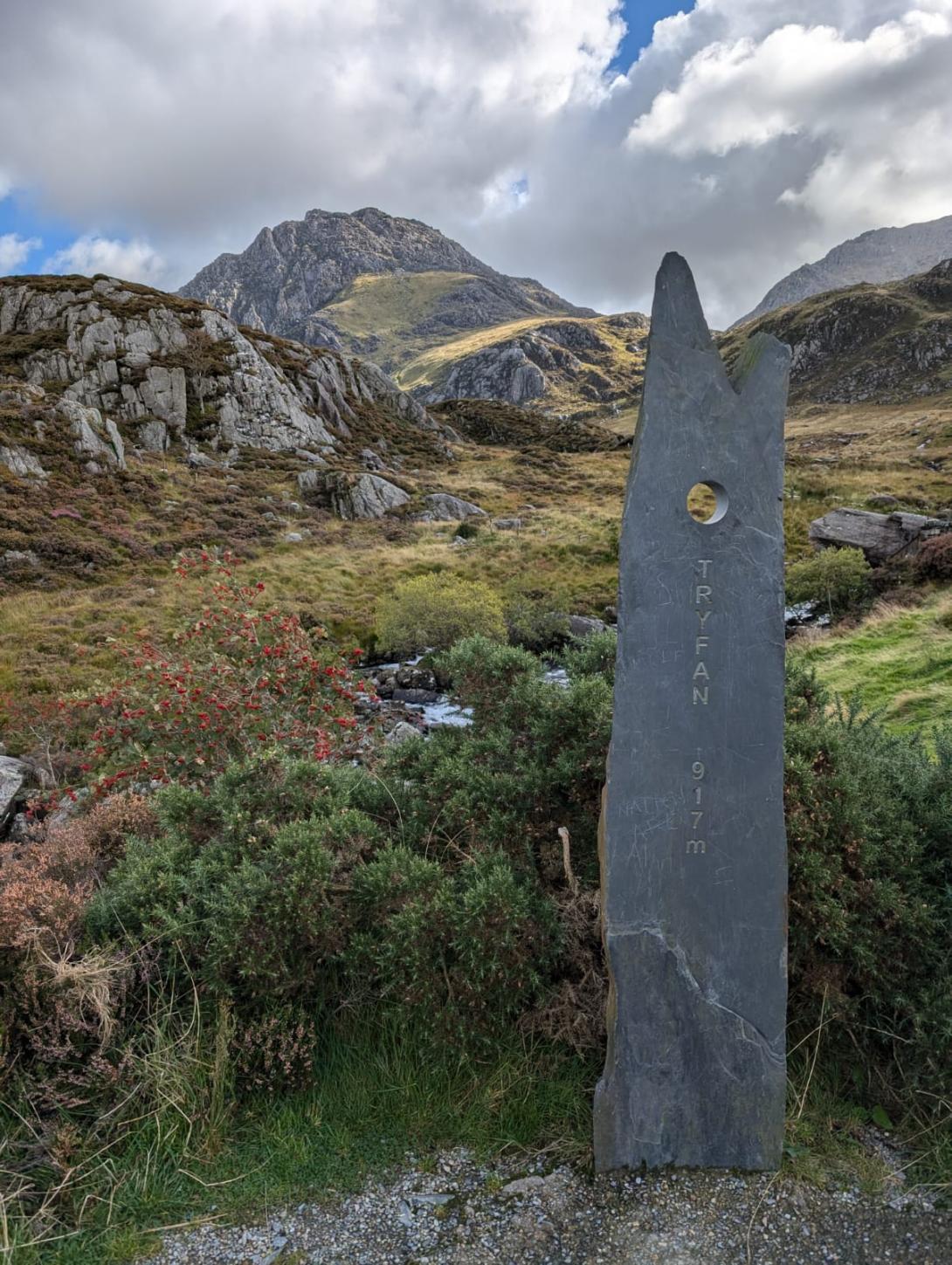 A tall, dark grey, jagged stone monument with a circular hole near the top. Text carved vertically down the stone reads 'TRYFAN 917m'. The monument stands on the side of a path with a stream and lush green and brown moorland to its left. In the background, a large, rugged mountain, called Tryfan, which rises under a sky with scattered clouds.