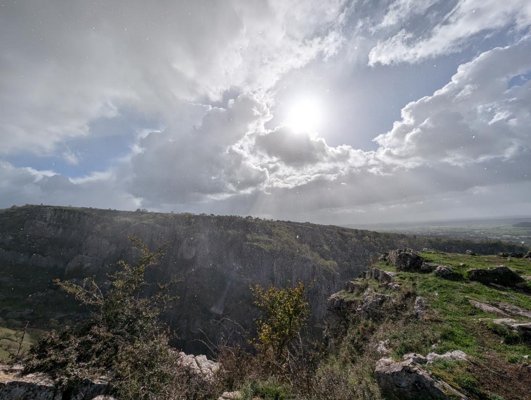 Dramatic view from the Cheddar Gorge Cliff-top Walk. The photograph captures the sheer, tree-lined cliff face of the gorge plunging down into the valley, seen from a rocky, grassy outcrop on the opposite side. The scene is bathed in sunlight breaking through high, bright clouds, creating visible sun rays and illuminating the fine mist and rain that speckles the air above the deep ravine.