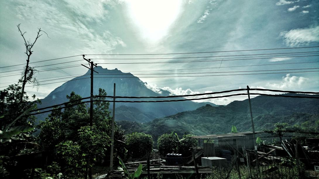A powerful view of the jagged peak of Mount Kinabalu emerging through the clouds, framed by power lines and lush green hills. Local buildings sit scattered in the foreground, showing the remote setting of the Borneo expedition.